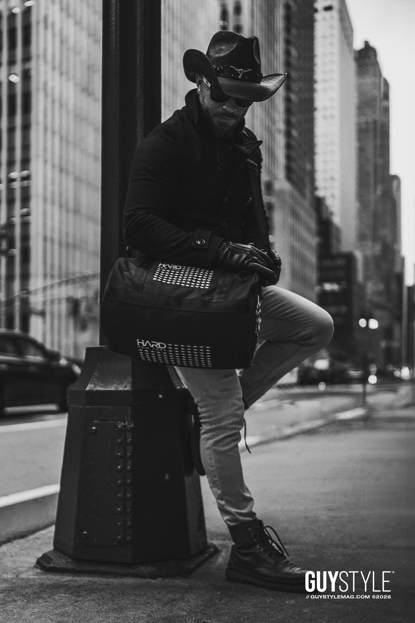 Maxwell Alexander leaning against a Midtown street post in a cowboy hat, black coat, pale skinny jeans and HARD NEW YORK bag.