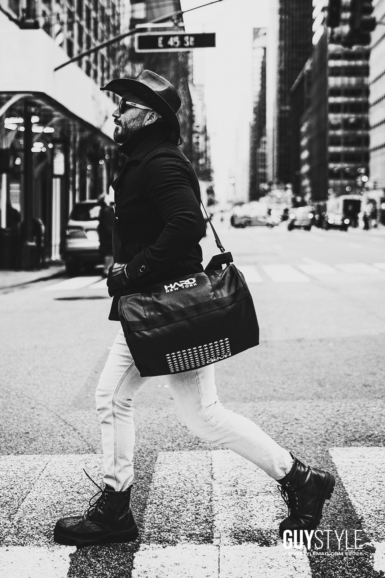 Maxwell Alexander striding across a Midtown Manhattan crosswalk in a cowboy hat, black coat, pale skinny jeans and HARD NEW YORK bag.