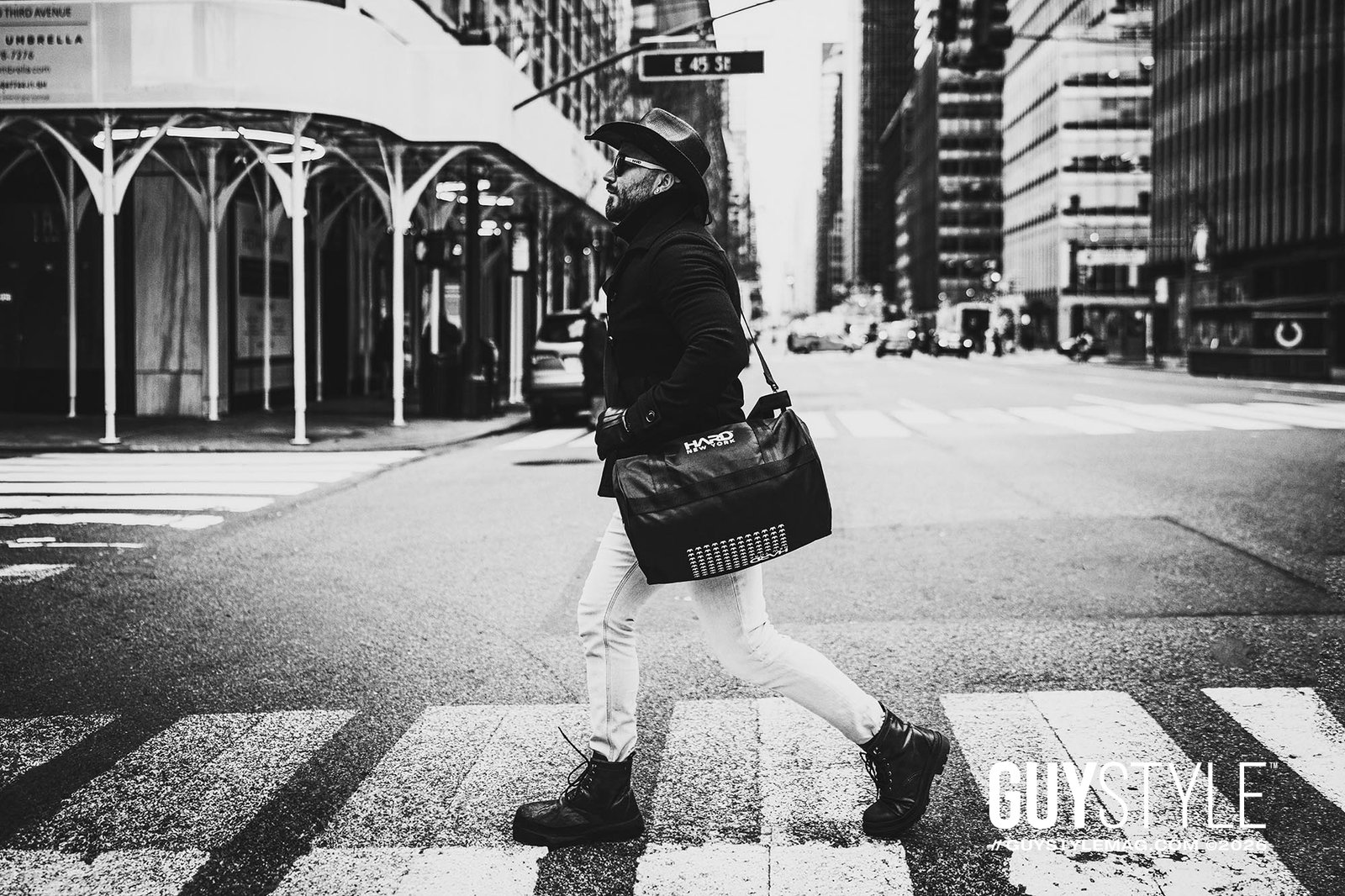 Maxwell Alexander crossing a Midtown Manhattan street in a cowboy hat, black coat, pale skinny jeans and HARD NEW YORK bag.