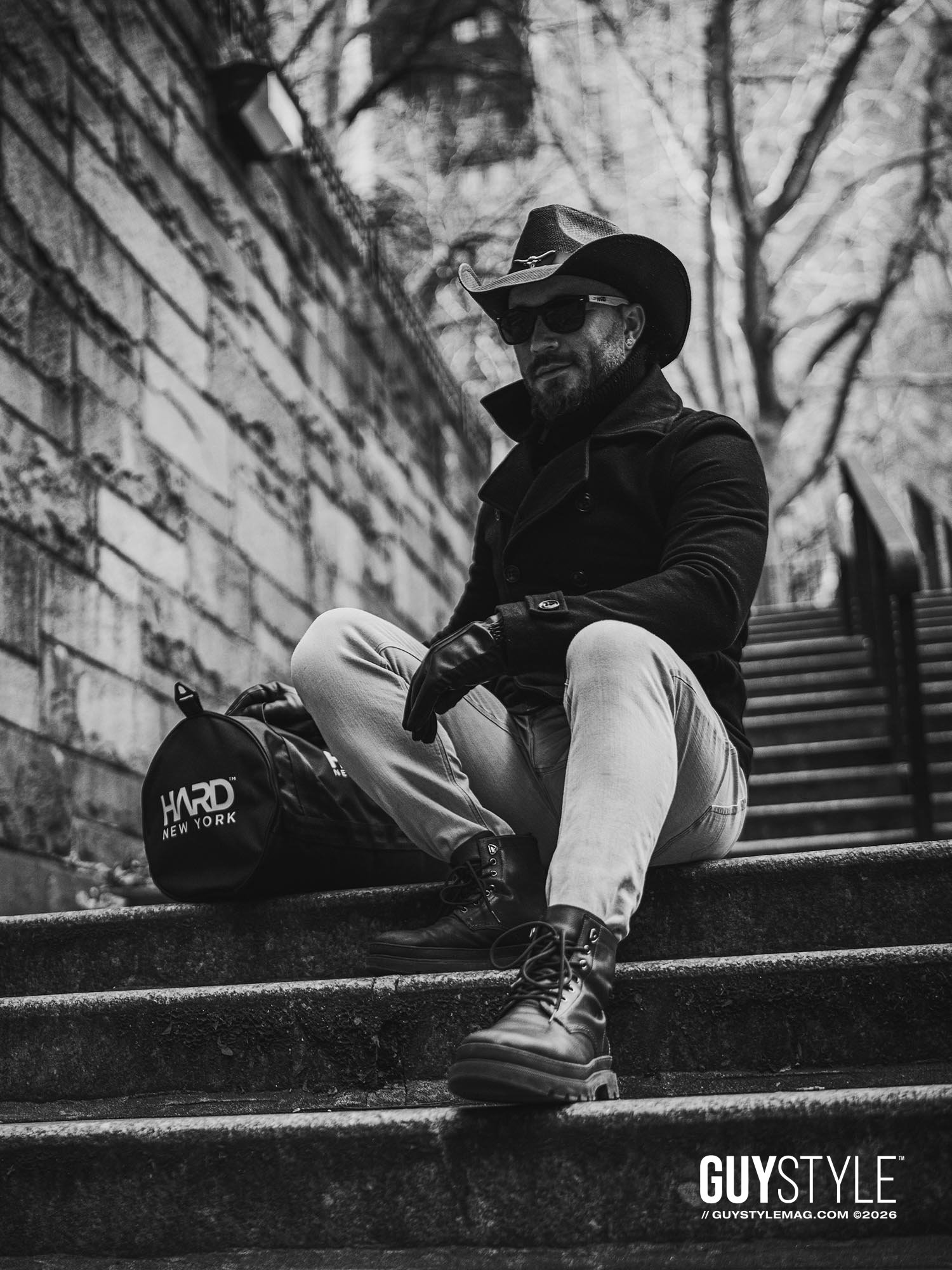 Maxwell Alexander seated on Tudor City steps in a cowboy hat, black coat, pale skinny jeans and combat boots beside a HARD NEW YORK bag.