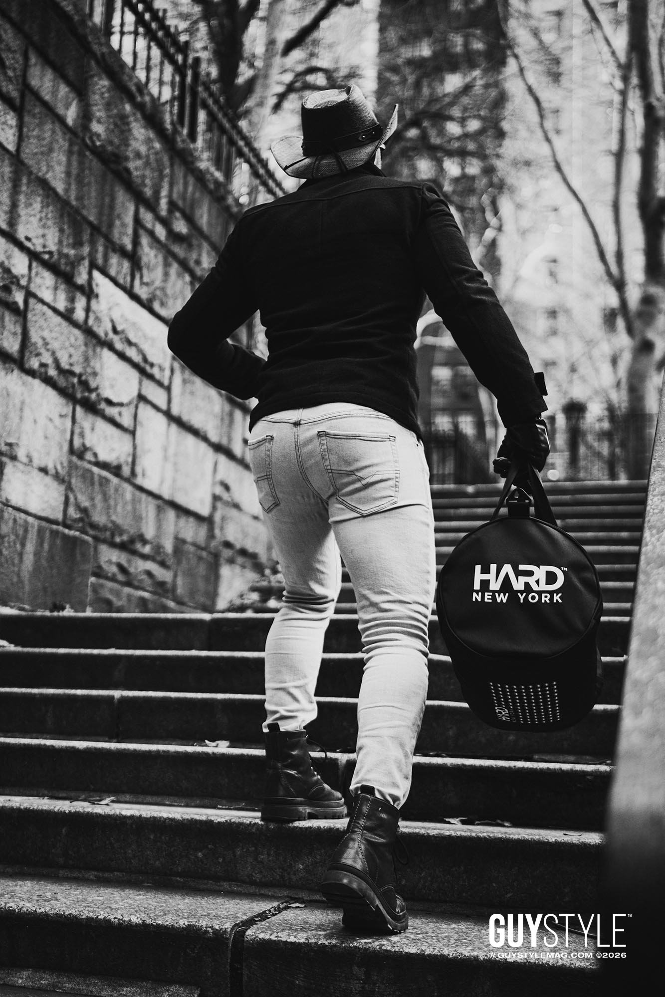 Maxwell Alexander walking up Tudor City steps in pale skinny jeans, black coat and cowboy hat while carrying a round HARD NEW YORK bag.