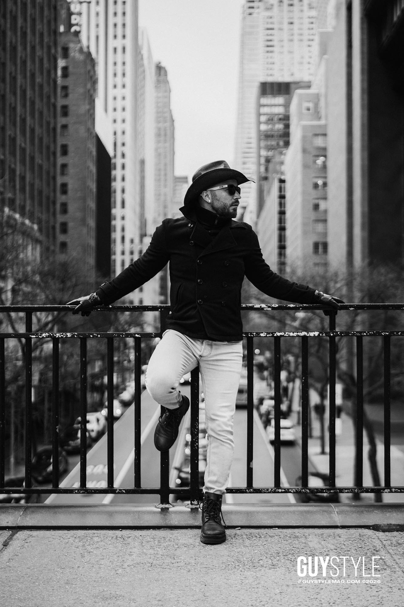 Maxwell Alexander posing above a Manhattan avenue in a cowboy hat, fitted black coat, pale skinny jeans and combat boots.