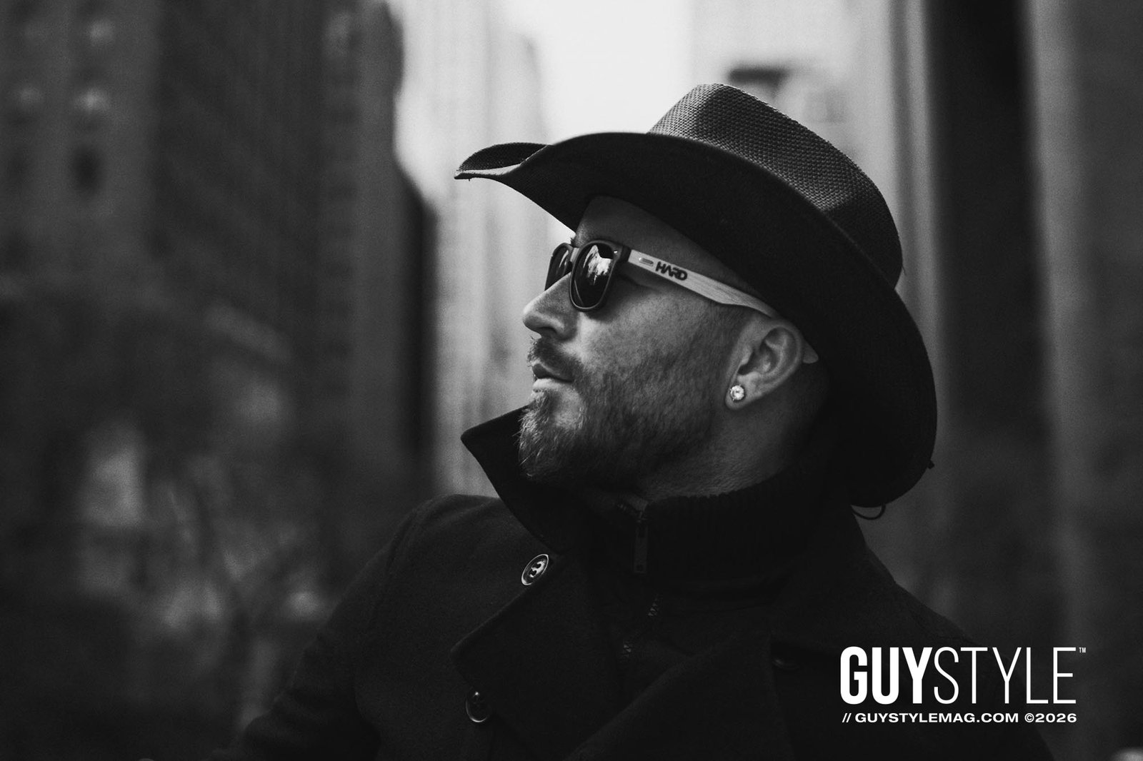 Profile portrait of Maxwell Alexander wearing a cowboy hat, sunglasses, black coat and diamond stud in Midtown Manhattan.