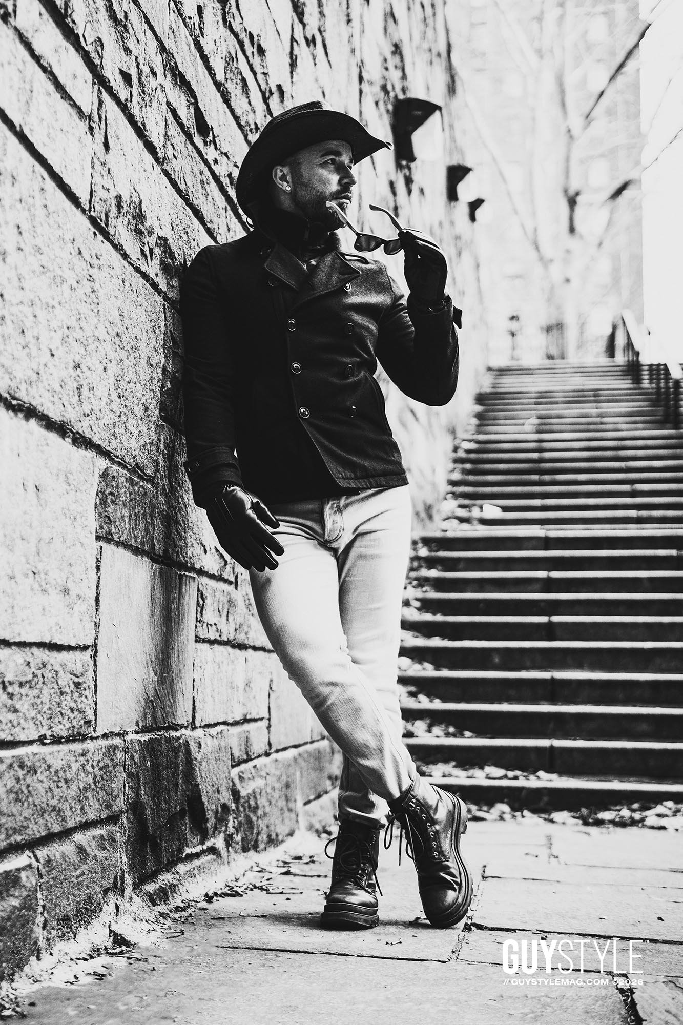 Maxwell Alexander posing against a stone wall in Tudor City wearing a cowboy hat, black coat, pale jeans and combat boots.