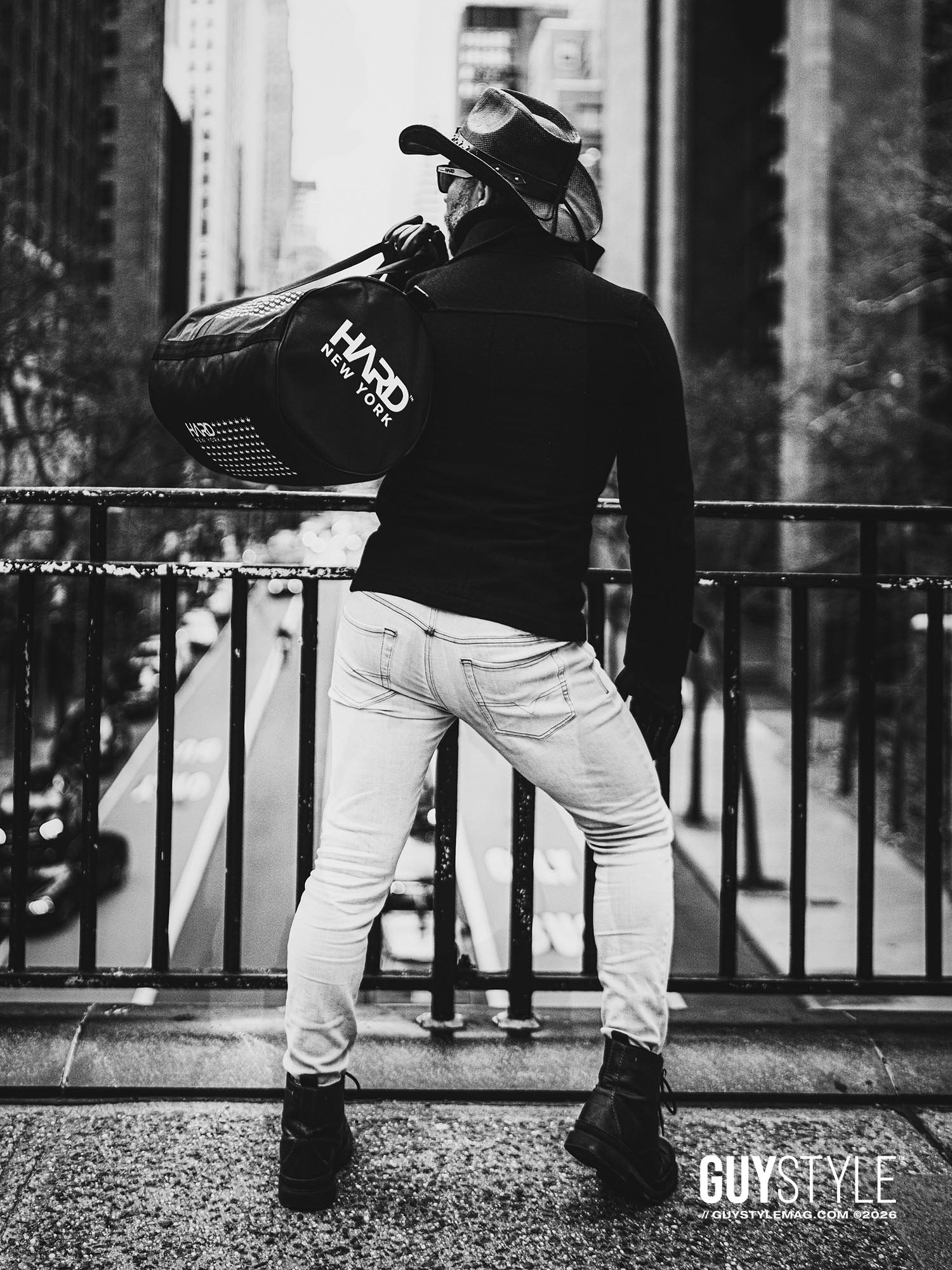 Rear view of Maxwell Alexander in pale skinny jeans and cowboy hat carrying a HARD NEW YORK bag above a Manhattan avenue.