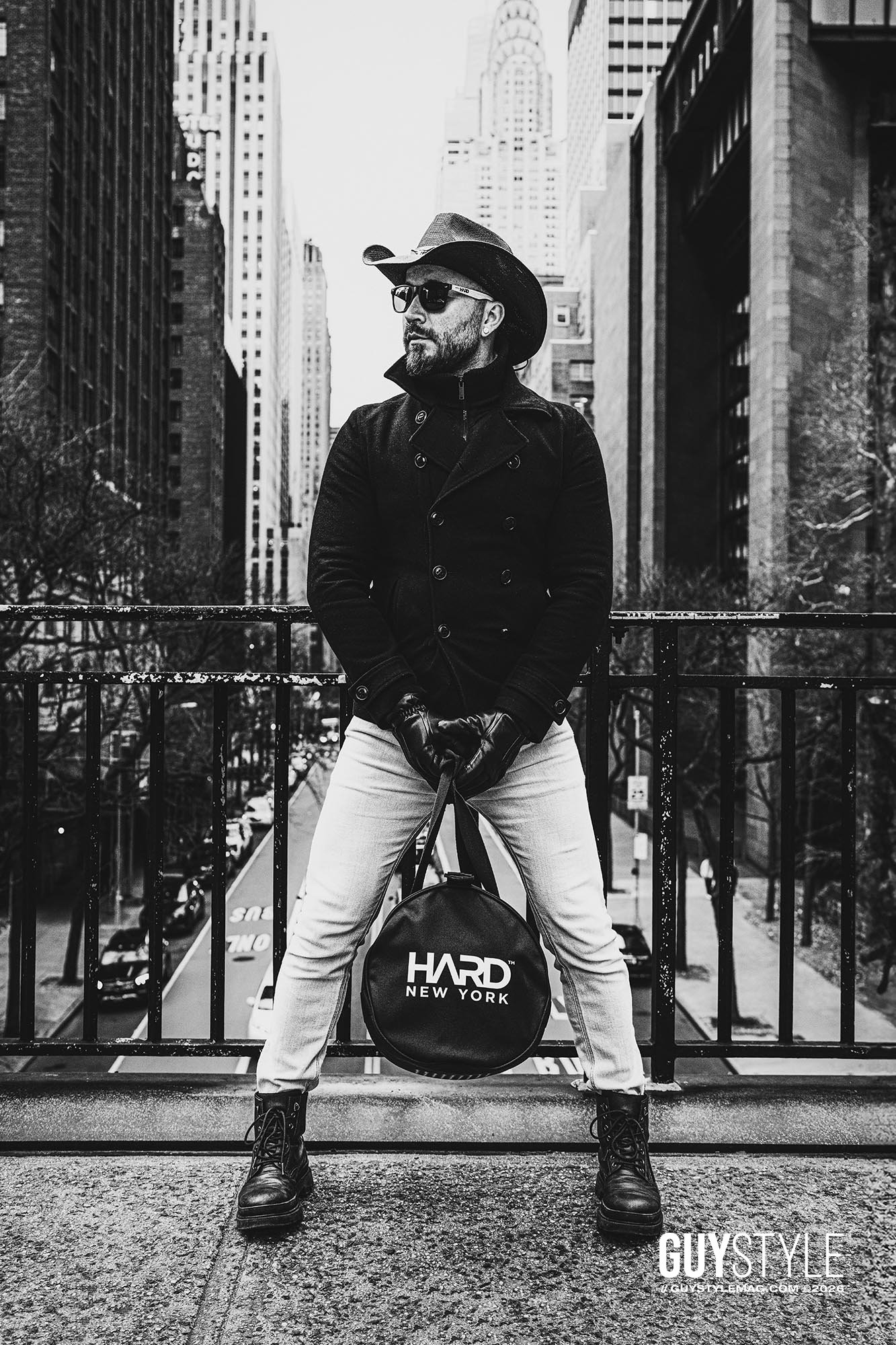 Maxwell Alexander standing above Manhattan in a cowboy hat and fitted black coat with a round HARD NEW YORK bag between his legs.
