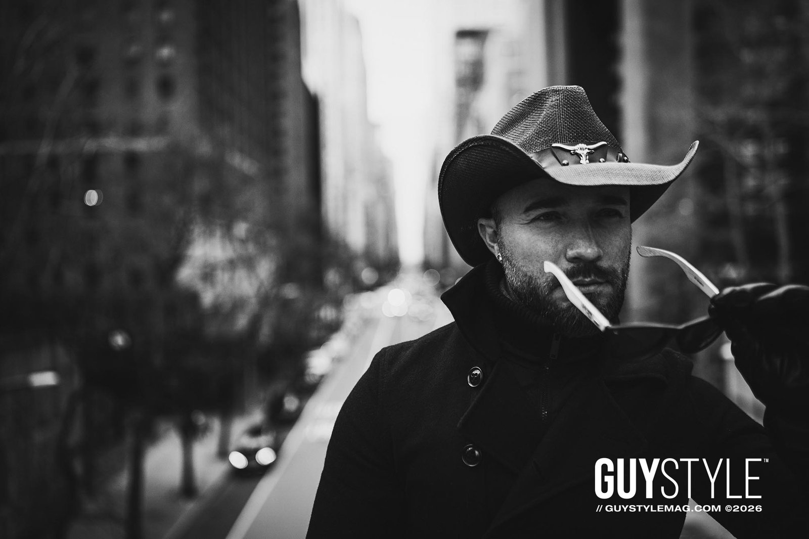 Black-and-white portrait of Maxwell Alexander in a cowboy hat, black coat and leather gloves holding sunglasses in Midtown Manhattan.